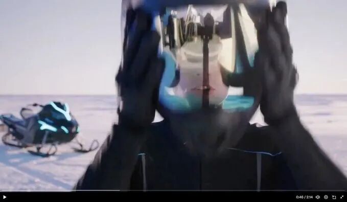 A person wearing a motorcycle helmet with a reflective visor, hands raised to the face, showing a distorted reflection of a camera. A snowmobile with blue lights is visible in the snowy background. A timestamp overlay at the bottom reads "04:24."