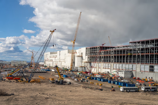 Construction of Building ID1 at Micron Boise, with cranes, heavy machinery, and partially built structures under a cloudy sky.