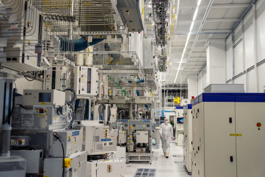 一名身穿全套无尘室服的人走在明亮的过道上，穿过一排排大型复杂机械，位于研发工厂中。