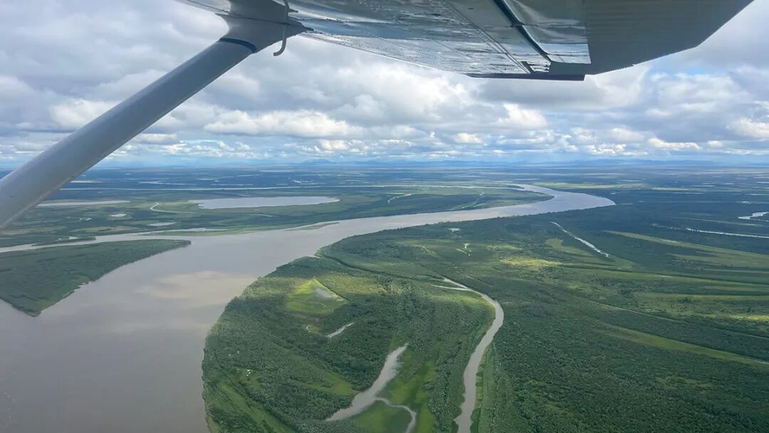 A river flowing through a lush green area is seen from a plane. The wing of the plane, as well as clouds, is visible at the top of the frame.